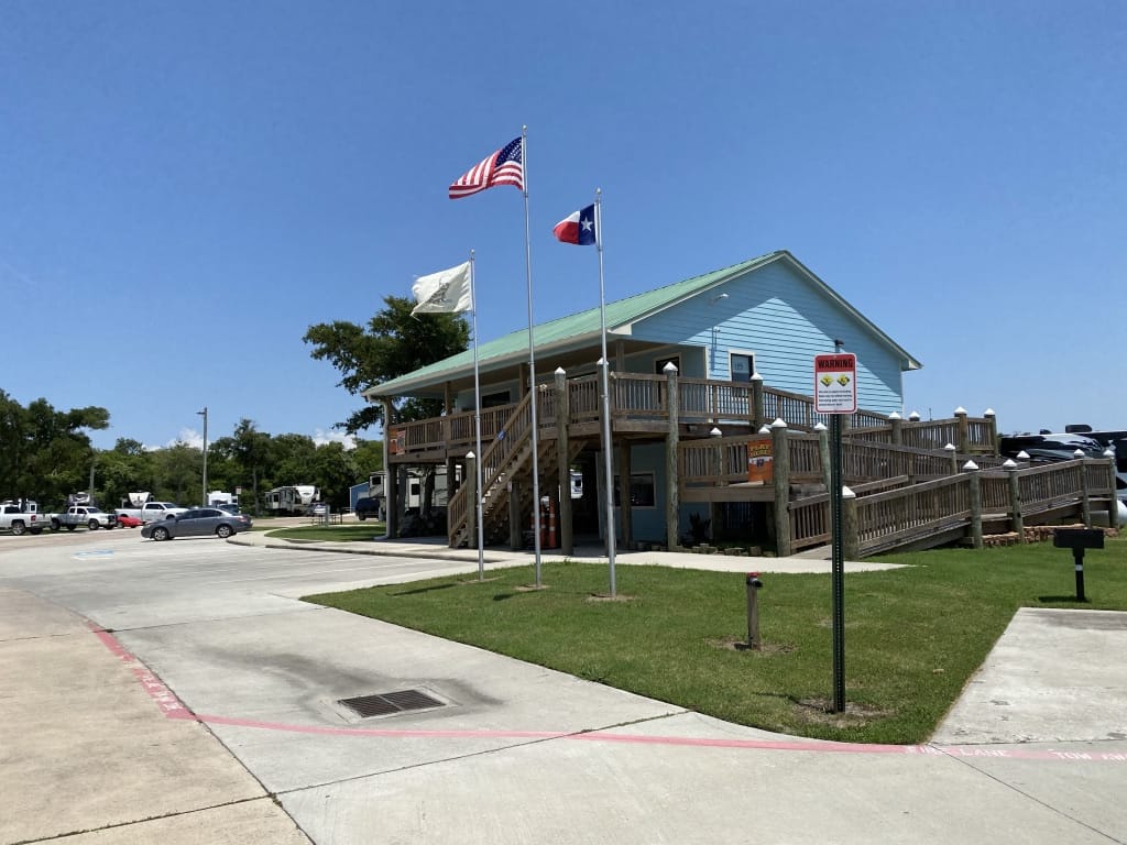 Sunny Day at Cedar Cove RV Resort A two-story building with a wooden staircase and multiple flags, including the United States and Texas flags, flying in front. The clear blue sky suggests a sunny day at Cedar Cove RV Resort.
